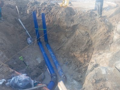 Wykop z dwoma niebieskimi rurami kanalizacyjnymi, połączonymi zielonym sznurkiem, widoczne narzędzia i fragment osoby w tle. Wykonane przyłącze wod-kan.