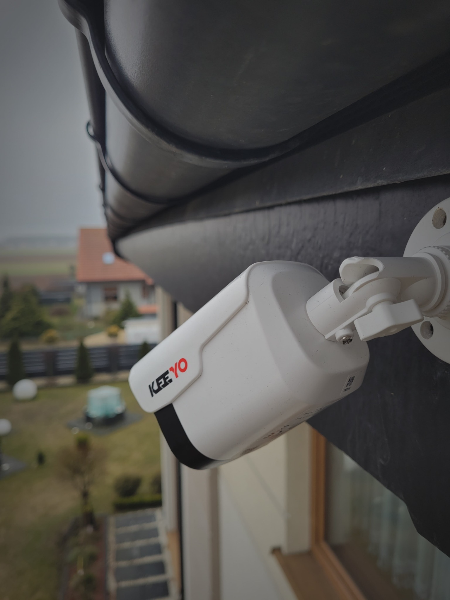 Biała kamera monitoringu marki KEEYO zamontowana na elewacji domu, z widokiem na ogród i okoliczne budynki w tle. Ujęcie z dołu, podkreślające perspektywę.