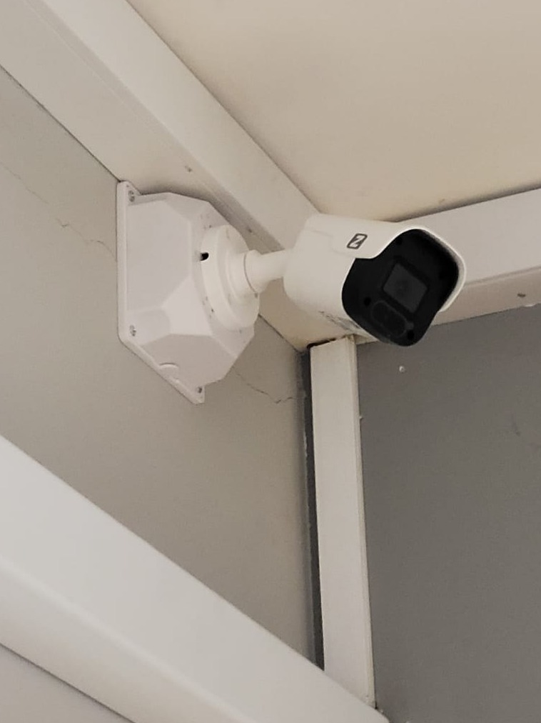 Biała kamera monitoringu, zamontowana na szarej ścianie w rogu pomieszczenia, z widoczną obudową i obiektywem skierowanym w dół.