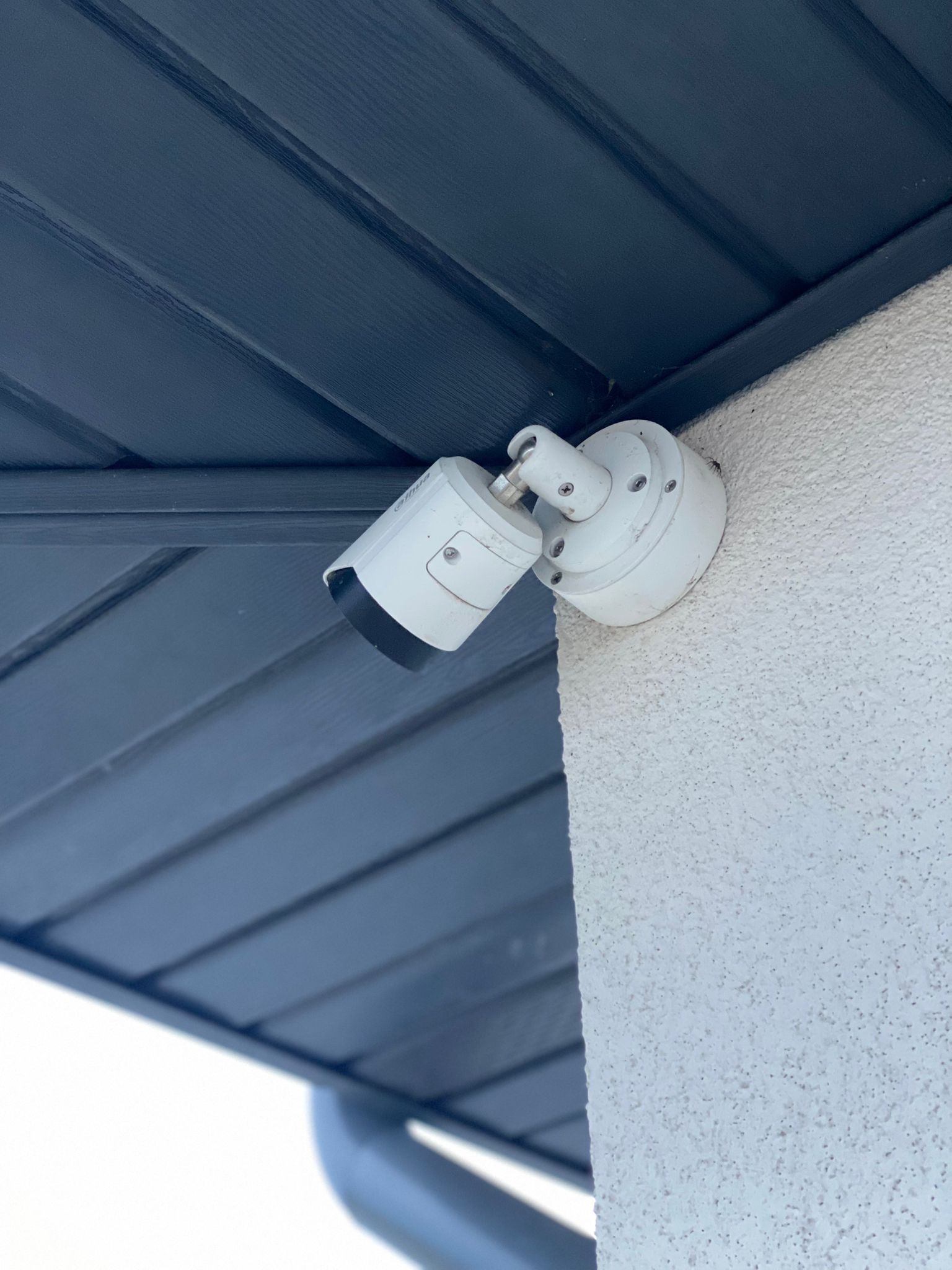 Biała kamera monitoringu przymocowana do ściany budynku z szorstkim tynkiem, widoczna spod ciemnej podbitki dachowej. Kąt ujęcia z dołu.