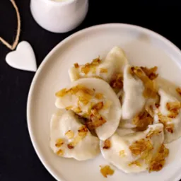 Apetyczne pierogi z karmelizowaną cebulką na białym talerzu, obok dzbanuszek ze śmietaną i dekoracja w kształcie serca na czarnym tle.