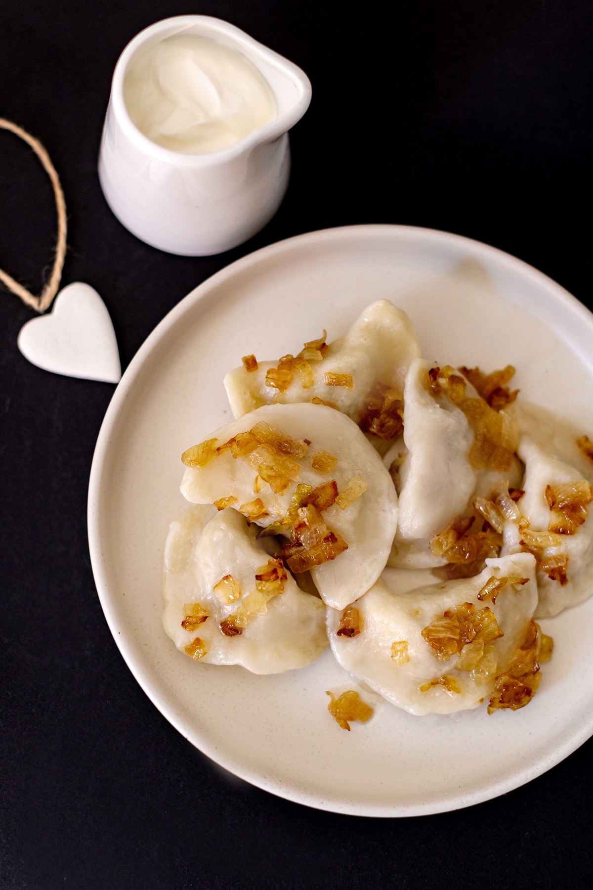 Apetyczne pierogi z karmelizowaną cebulką na białym talerzu, obok dzbanuszek ze śmietaną i dekoracja w kształcie serca na czarnym tle.