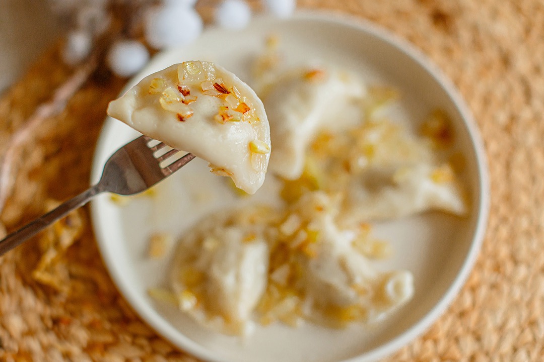 Pierogi z cebulką na widelcu, zbliżenie na farsz. Na talerzu obok kolejne pierogi, całość na plecionym, beżowym podkładzie. Delikatne, naturalne światło.