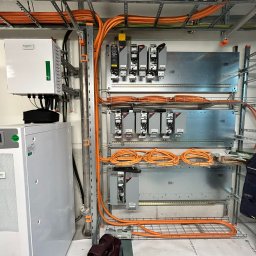Jacek Goły - Rozbudowana instalacja elektryczna w budynku, z pomarańczowymi kablami ułożonymi na metalowych półkach i szafach sterowniczych Schneider Electric.