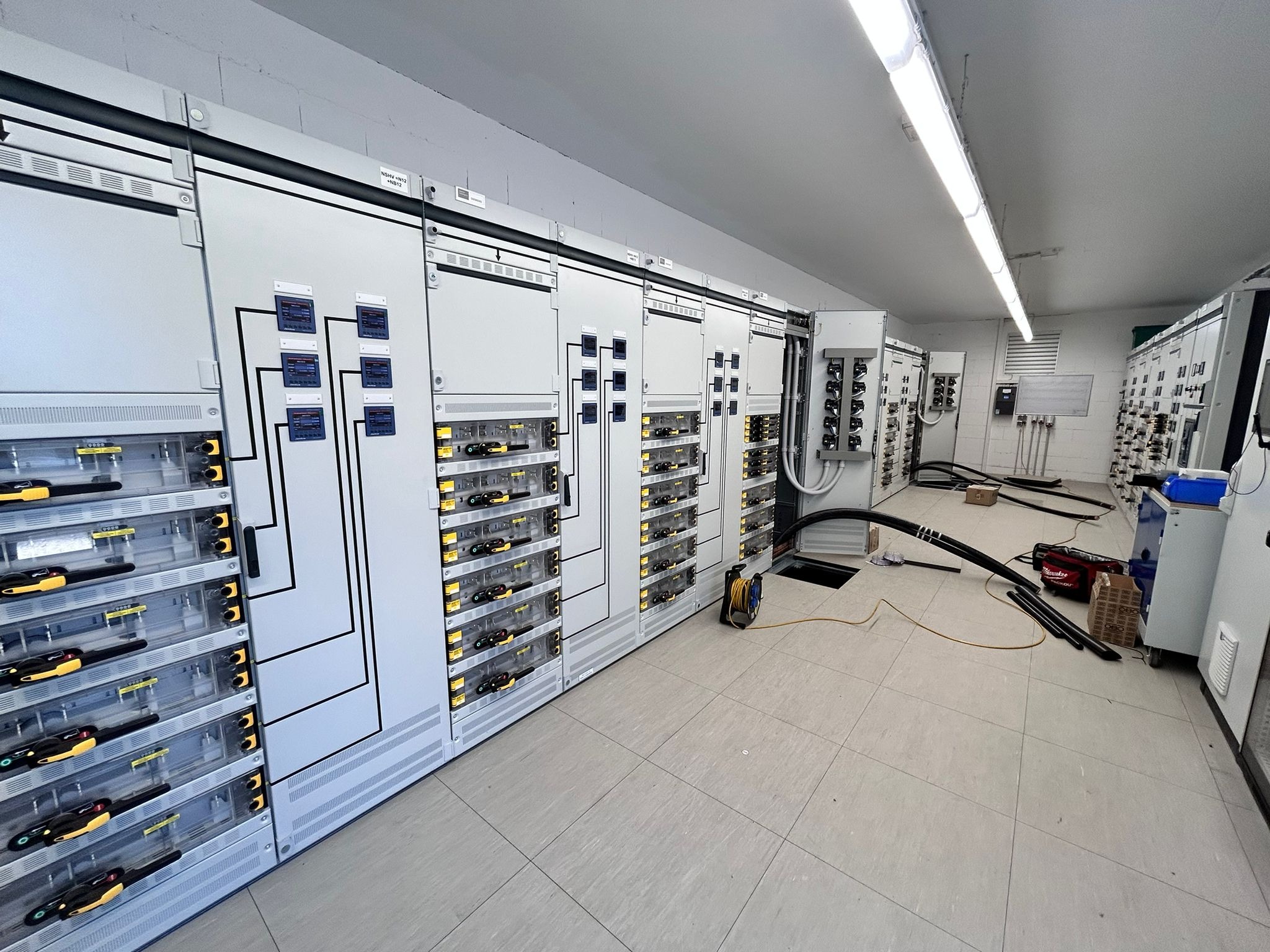 Duża rozdzielnica elektryczna w pomieszczeniu technicznym z oprzyrządowaniem. Widoczne kable, narzędzia i elementy sterujące.