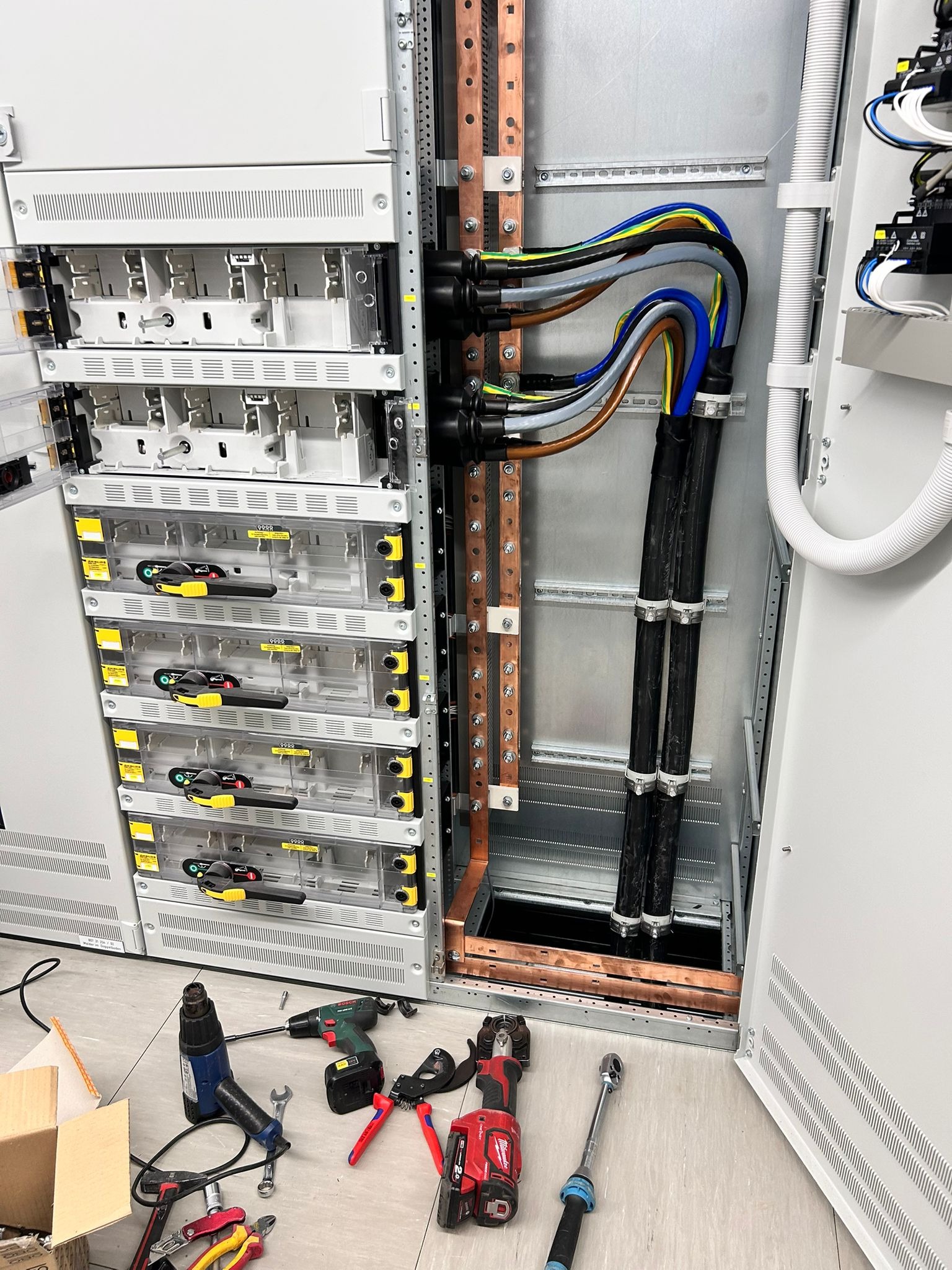 Rozdzielnia elektryczna w trakcie montażu: widoczne kable, szyny miedziane, zabezpieczenia i narzędzia na podłodze. Detal prac instalacyjnych w skrzynce elektrycznej.