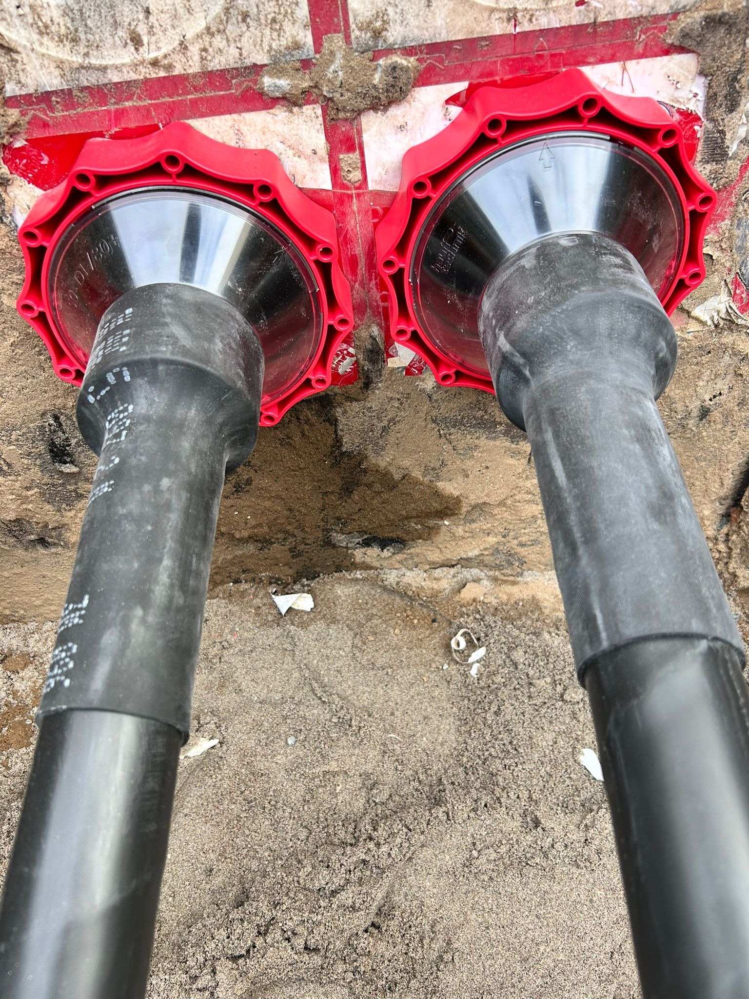 Dwa czarne kable wchodzące do czerwonych, okrągłych złączek na piasku. Widok z bliska na etapie instalacji.