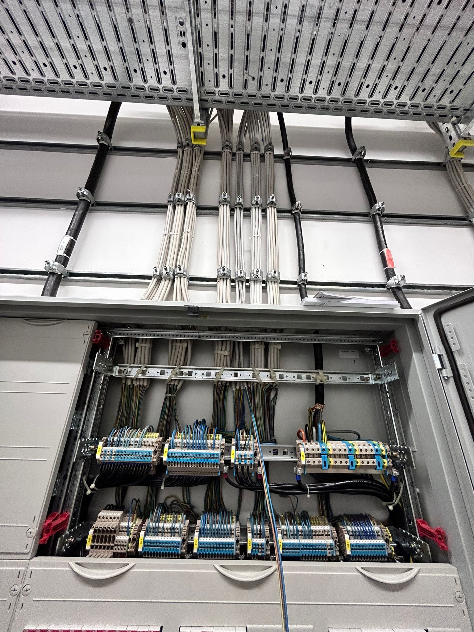 Otwarta skrzynka rozdzielcza z licznymi przewodami i złączkami wewnątrz. Widoczne kable prowadzone w korytach kablowych pod sufitem.