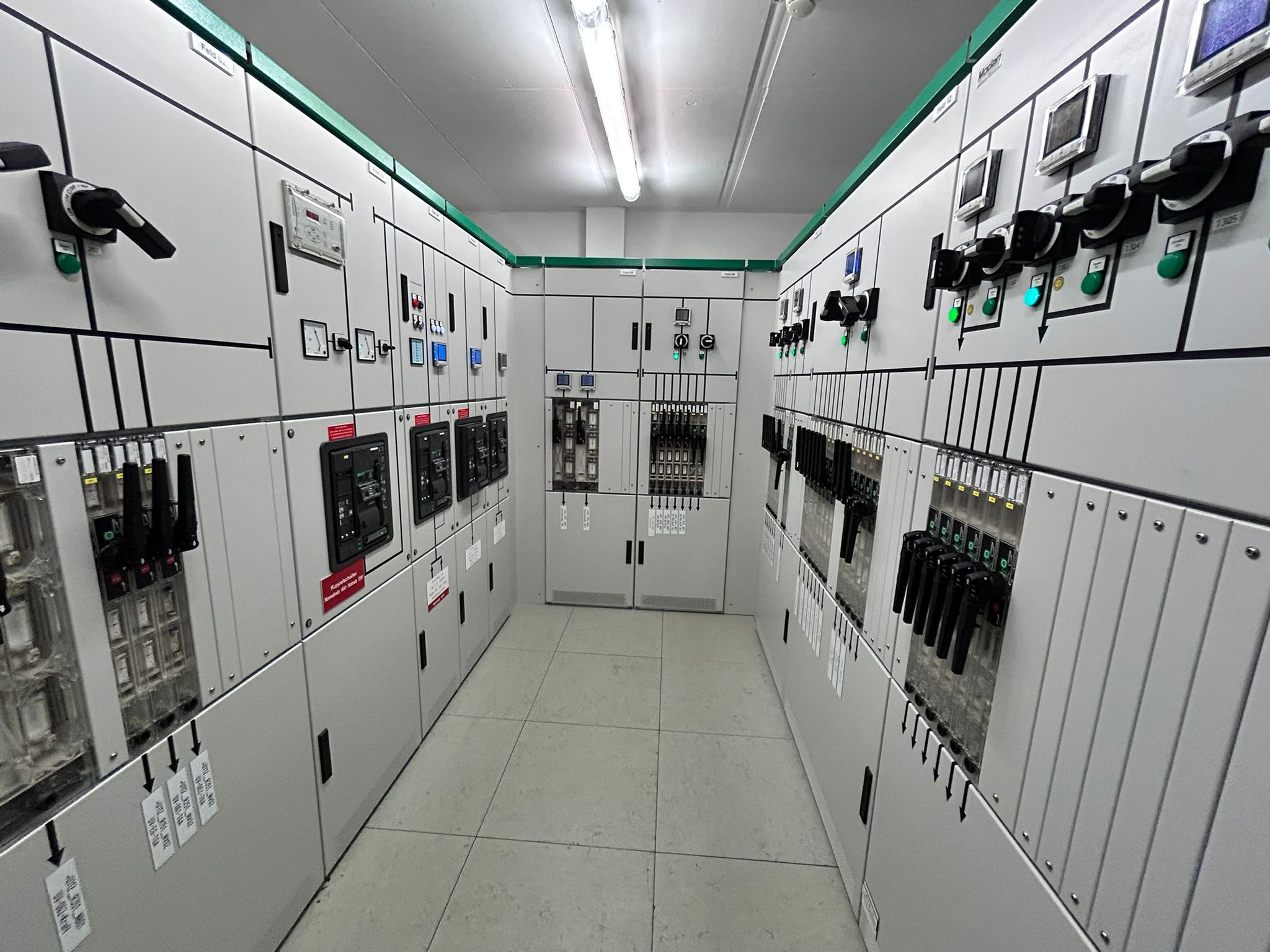 Rozdzielnica elektryczna z licznymi przełącznikami i wskaźnikami, widok z perspektywy korytarza w pomieszczeniu technicznym z białymi panelami.