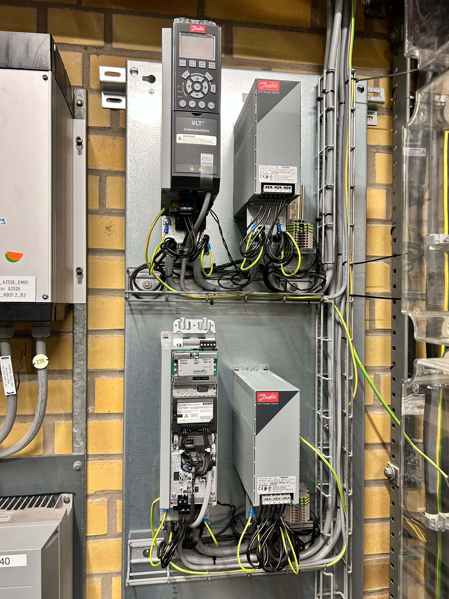 Rozdzielnica elektryczna Danfoss VLT z okablowaniem na ceglanej ścianie. Widoczne przewody, sterowniki i moduły zasilania. Element instalacji w szynie montażowej.