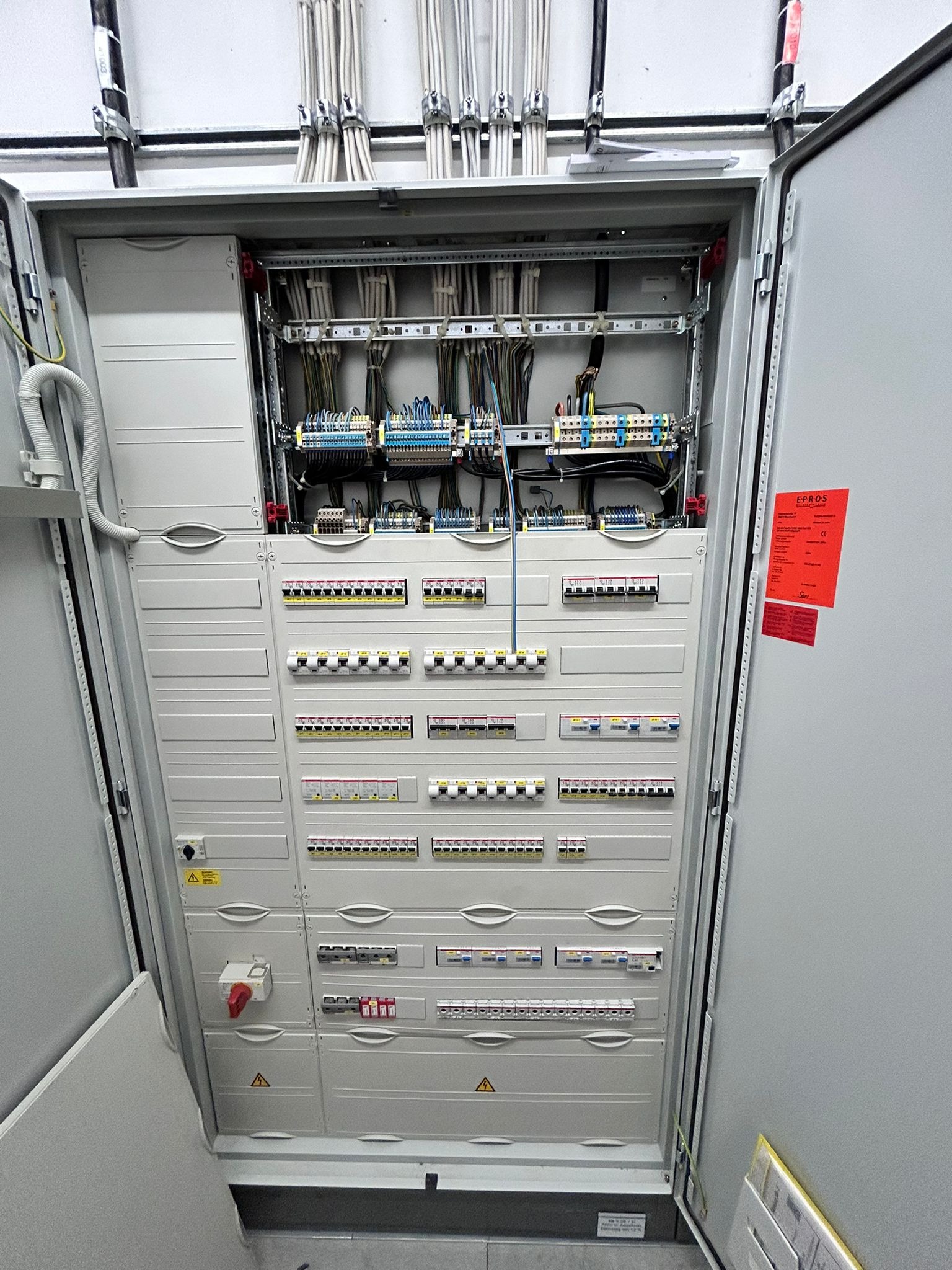 Otwarta, szara szafa elektryczna z widocznymi bezpiecznikami, przewodami i elementami sterującymi. W tle biała ściana i kable. Widoczne oznaczenia ostrzegawcze.