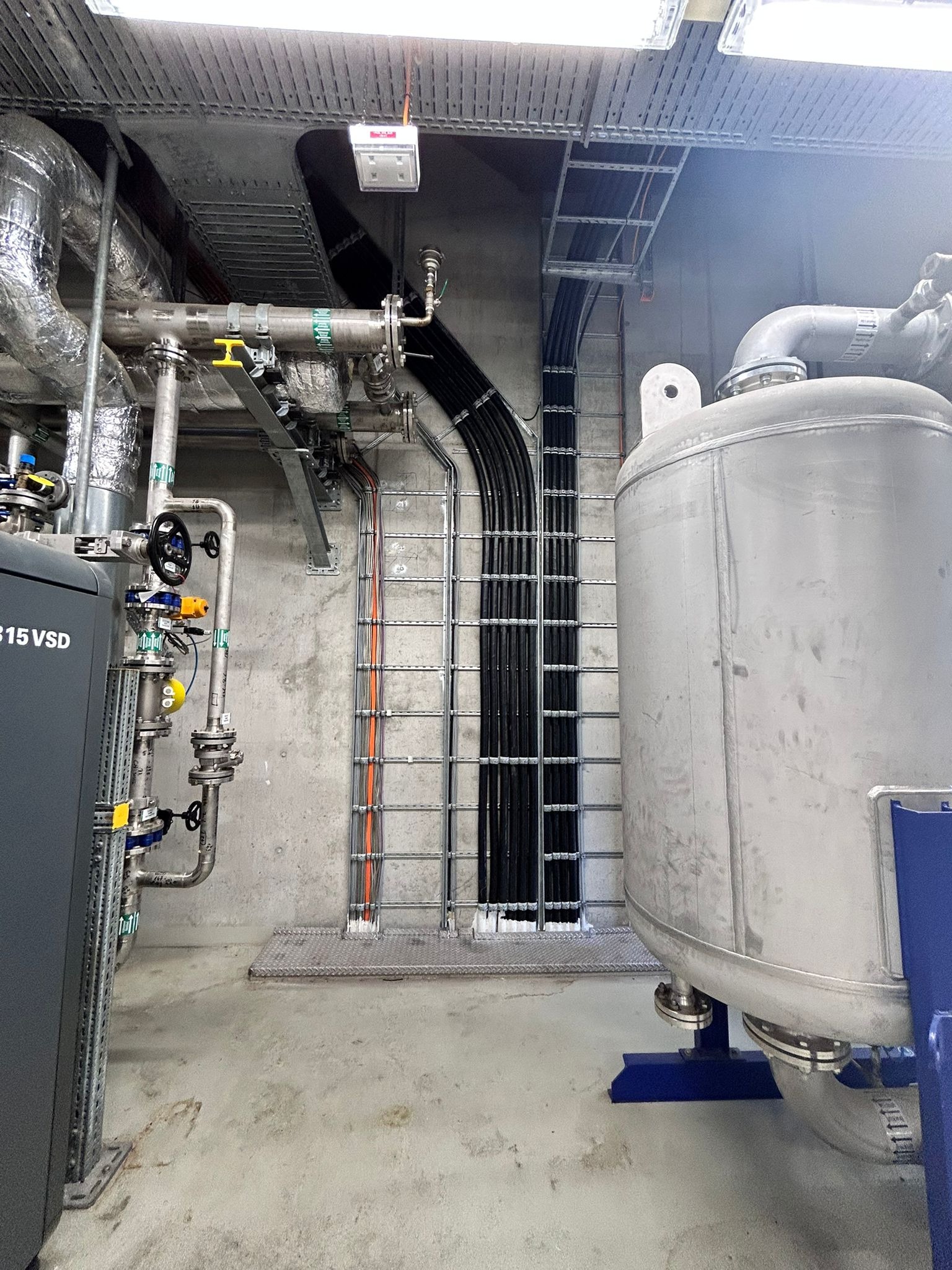 Kable w korytach i rury w surowym wnętrzu technicznym. Widoczne metalowe drabinki kablowe i orurowanie. Szara betonowa ściana i podłoga.