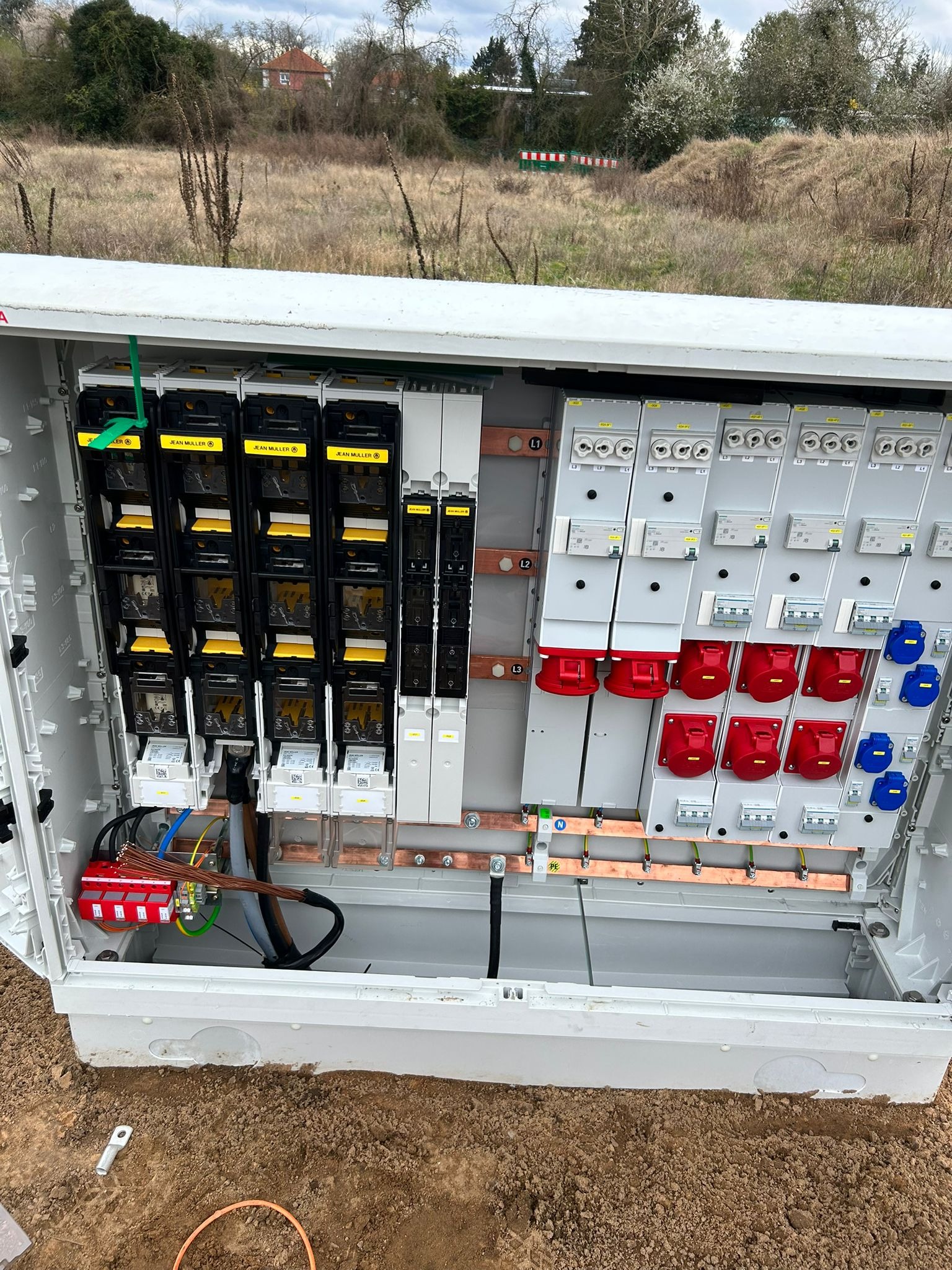 Otwarta skrzynka elektryczna z bezpiecznikami i gniazdami, widoczne okablowanie i elementy instalacji, tło stanowią drzewa i krzewy.