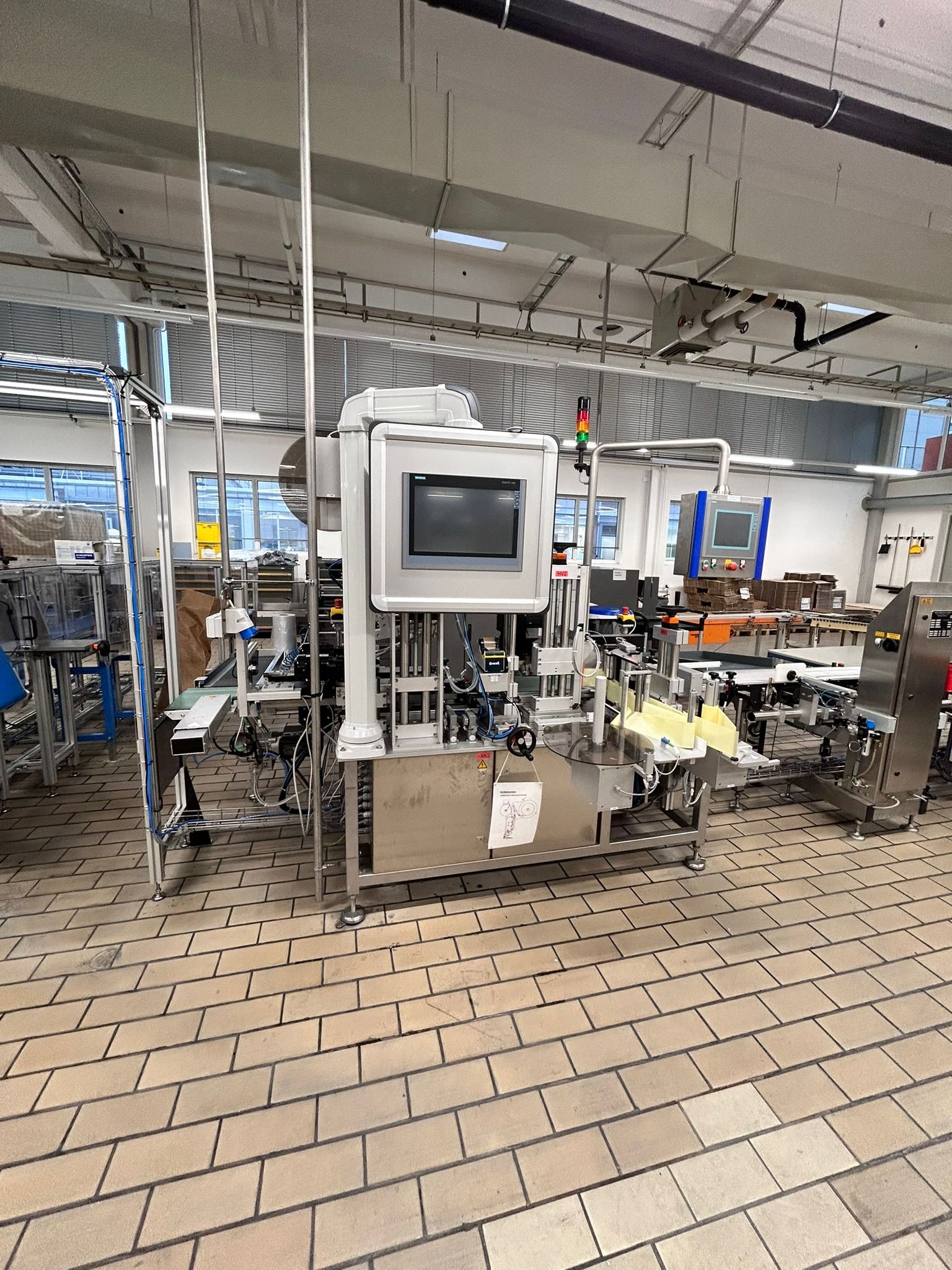 Automatyczna linia produkcyjna z panelami sterowania, zainstalowana w jasnym pomieszczeniu z kafelkową podłogą, widoczne rury i przewody.