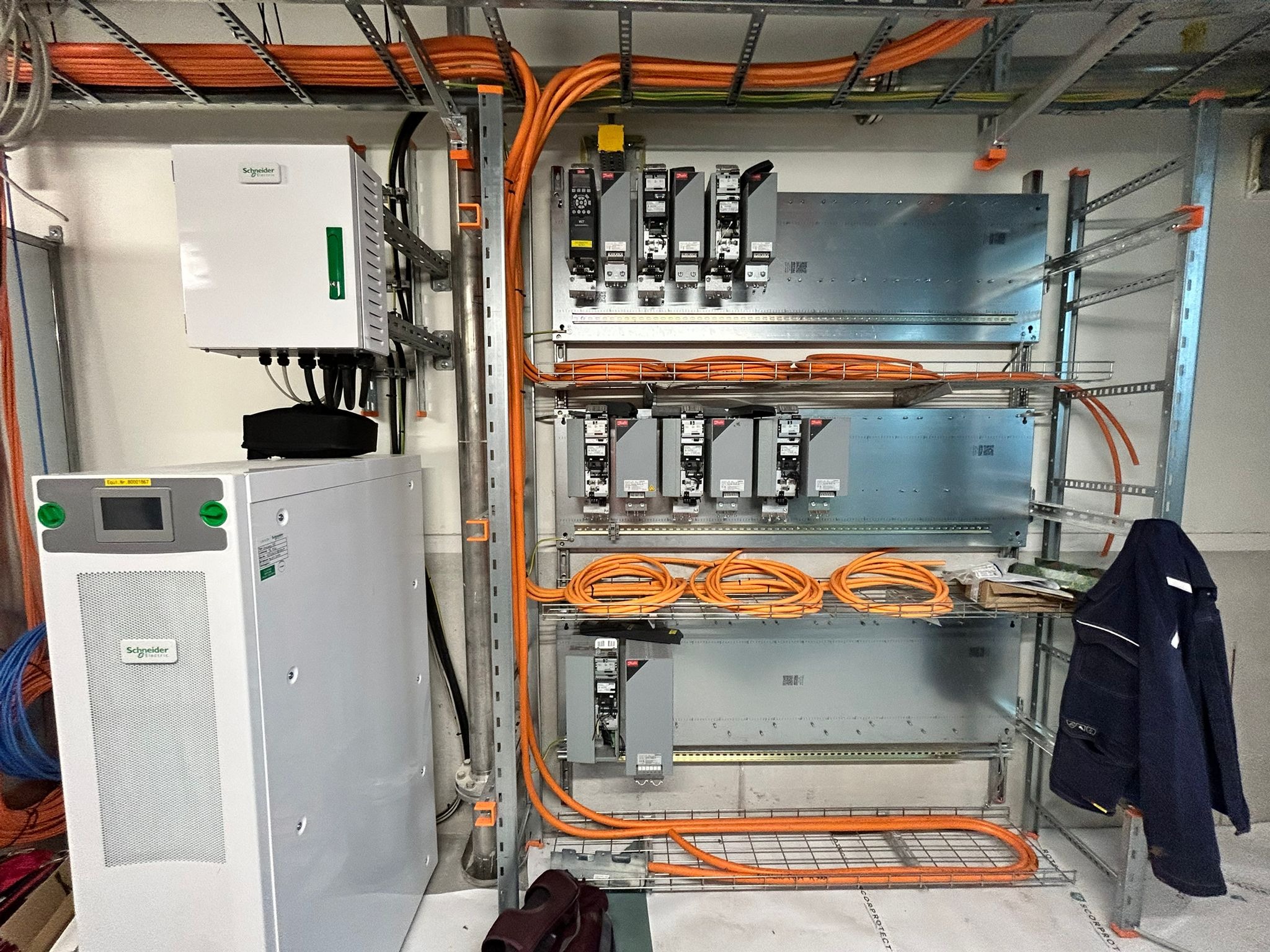 Rozbudowana instalacja elektryczna w budynku, z pomarańczowymi kablami ułożonymi na metalowych półkach i szafach sterowniczych Schneider Electric.