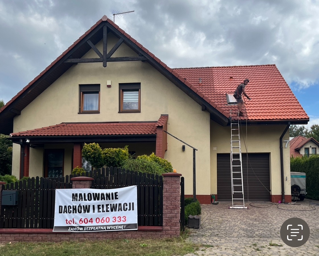 Pracownik na drabinie czyści dach domu z czerwonej dachówki. Na płocie baner z napisem 'Malowanie dachów i elewacji'.