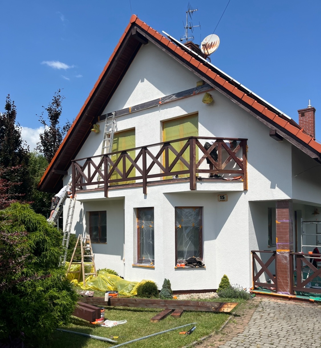 Biały dom z balkonem w trakcie odświeżania elewacji. Okna i elementy drewniane zabezpieczone folią, widoczne drabiny i narzędzia malarskie, fragment ogrodu.