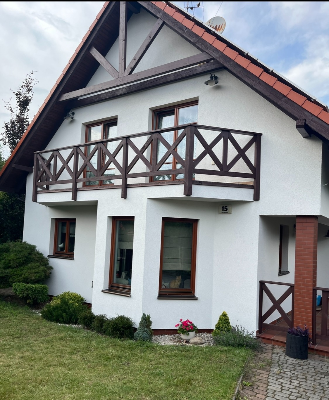 Biała elewacja domu z drewnianą konstrukcją dachu i balkonu. W oknie widoczny kot. Zadbane otoczenie z roślinami. Ujęcie z poziomu trawnika.