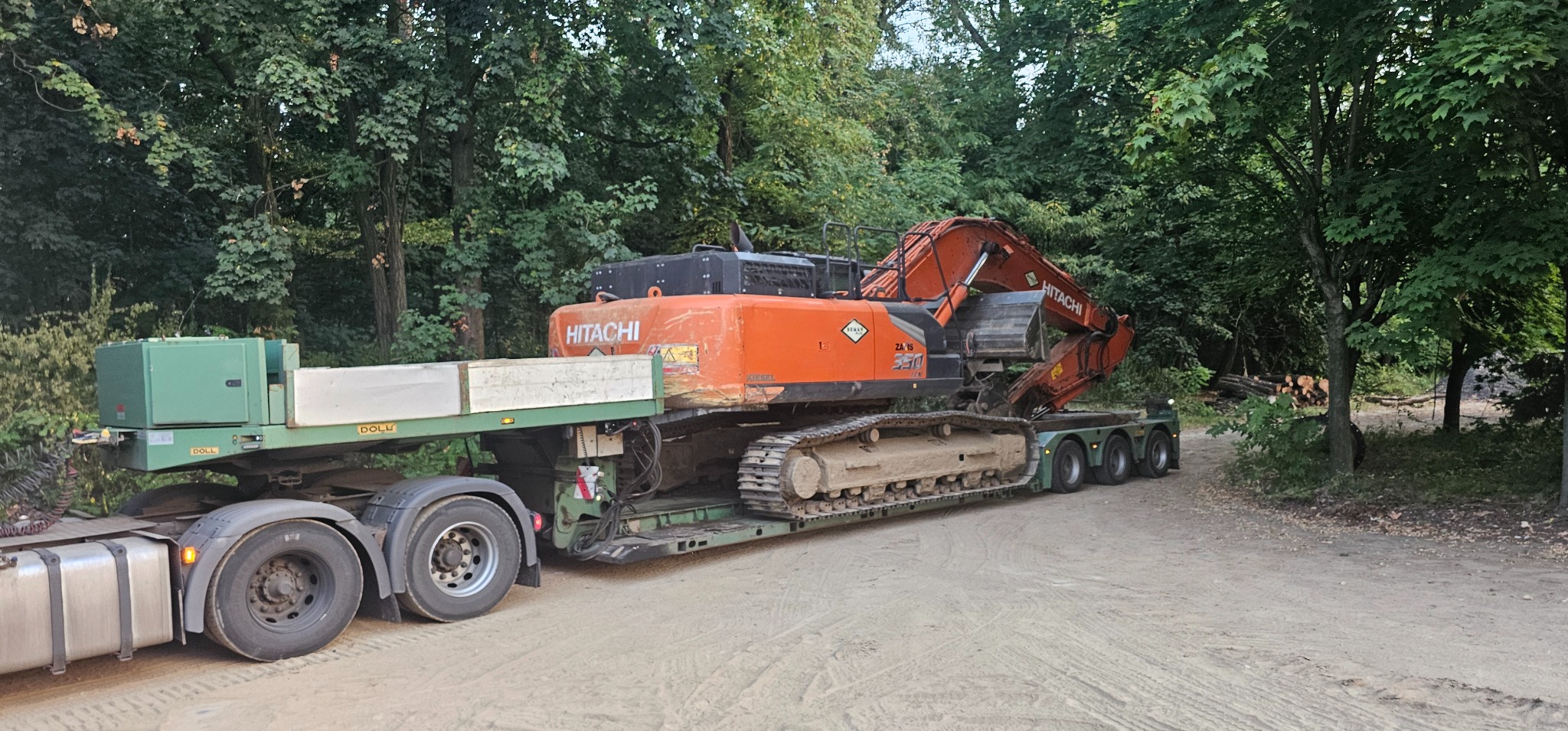 Pomarańczowa koparka Hitachi na niskopodwoziowej naczepie, transportowana po piaszczystej drodze w otoczeniu drzew. Widoczny fragment lasu w tle.