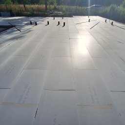 Krzysztof Wiśniewski - Ułożone płyty izolacyjne XPS na przygotowanym podłożu, widoczne wystające rury i pręty zbrojeniowe, teren budowy w tle.