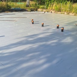 Krzysztof Wiśniewski - Izolacja fundamentów z płyt XPS, widoczne pionowe rury instalacyjne. Przygotowanie do wylewki, teren budowy w otoczeniu zieleni.