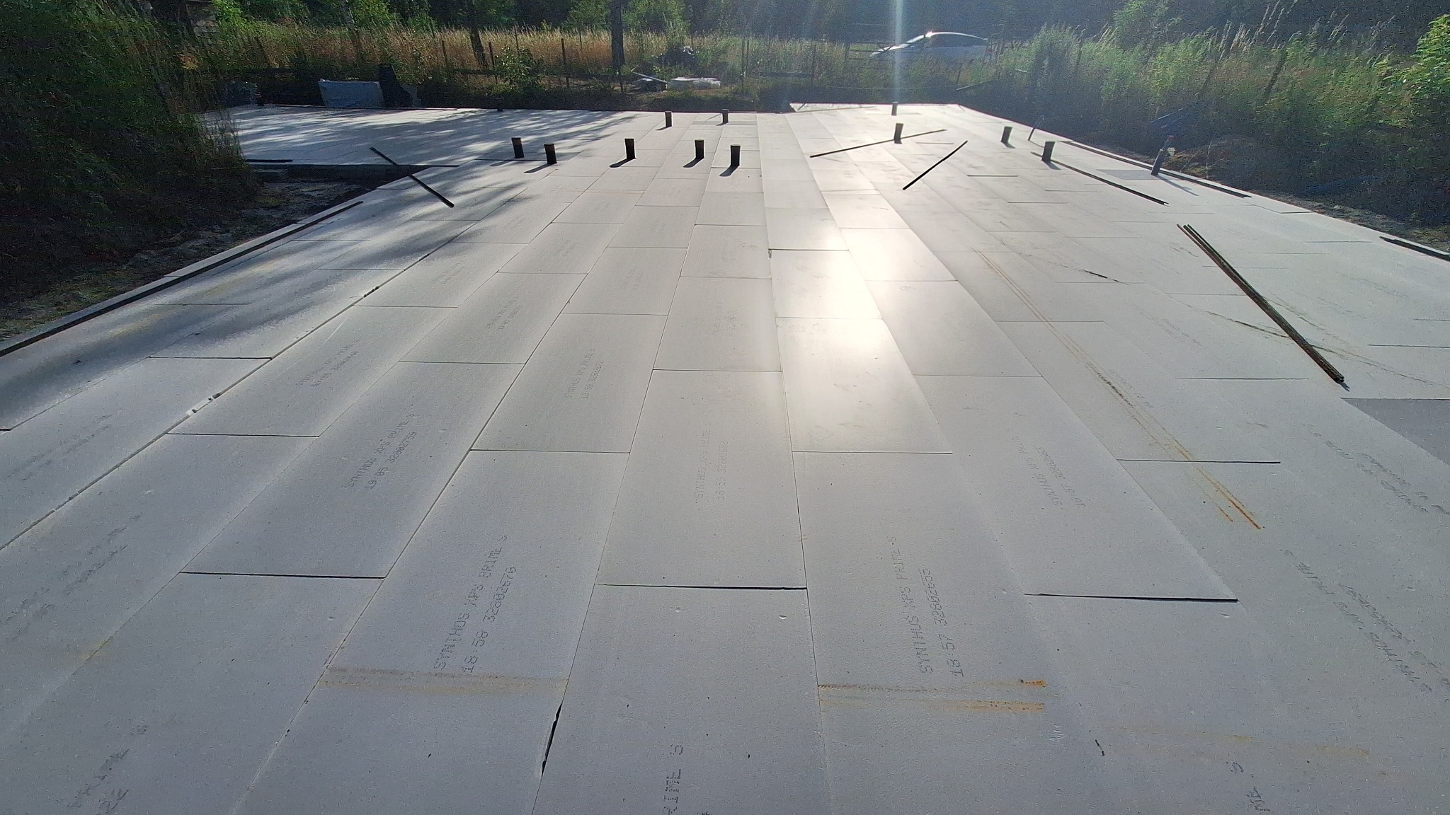 Ułożone płyty izolacyjne XPS na przygotowanym podłożu, widoczne wystające rury i pręty zbrojeniowe, teren budowy w tle.