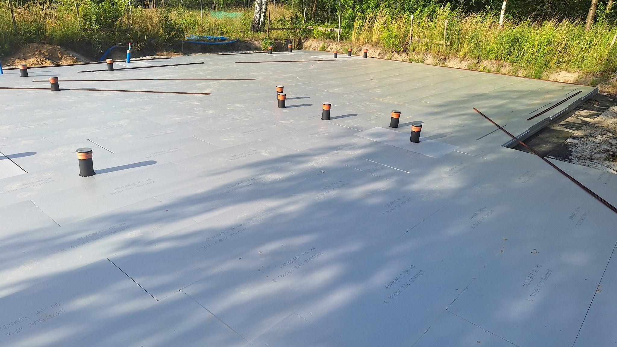 Izolacja fundamentów z płyt XPS, widoczne pionowe rury instalacyjne. Przygotowanie do wylewki, teren budowy w otoczeniu zieleni.