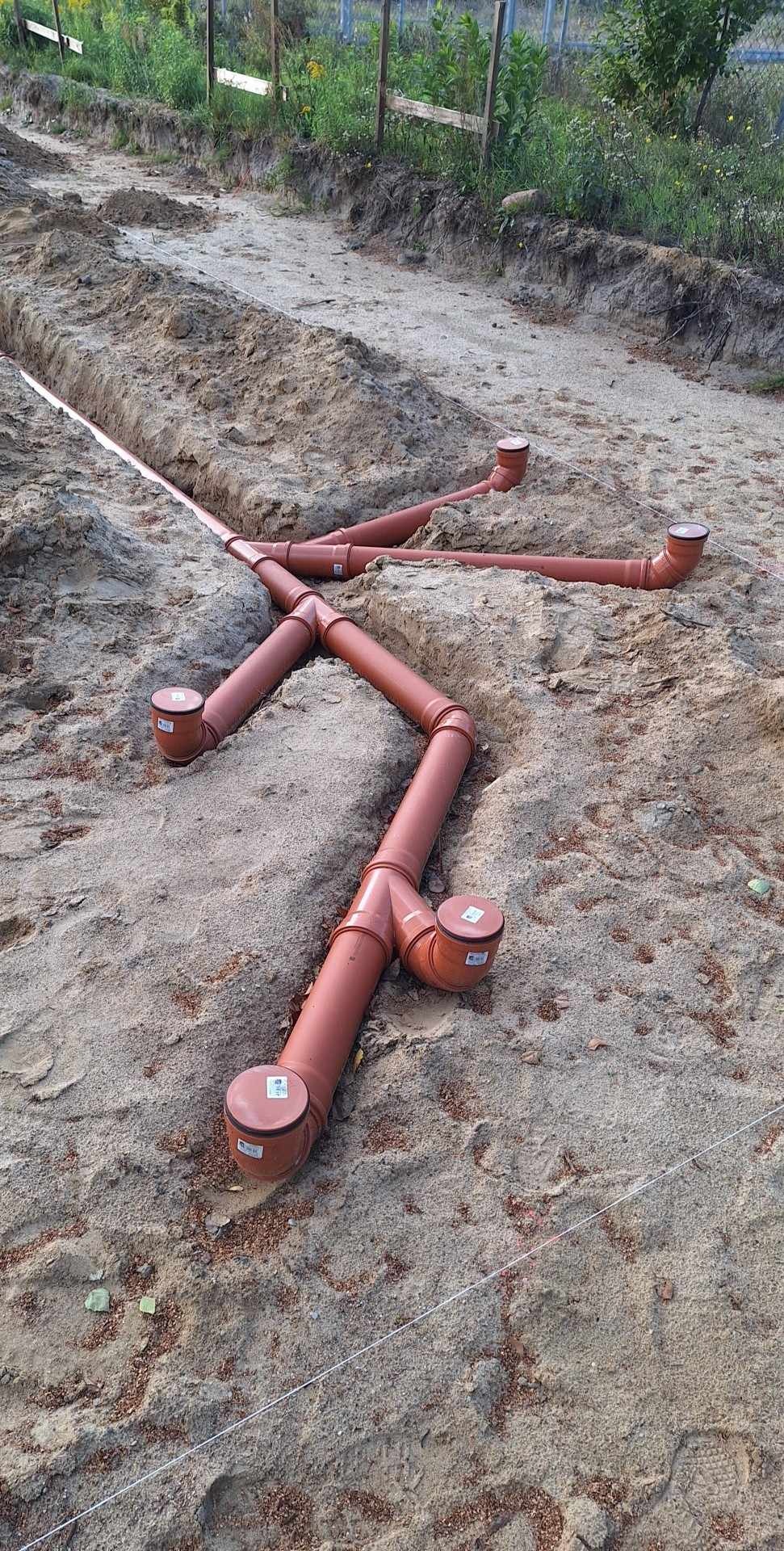 Brązowe rury kanalizacyjne ułożone w wykopie z piasku, widoczne z góry. Rury z uszczelkami i zaślepkami, częściowo zakopane w ziemi. Widoczne ślady stóp w piasku.