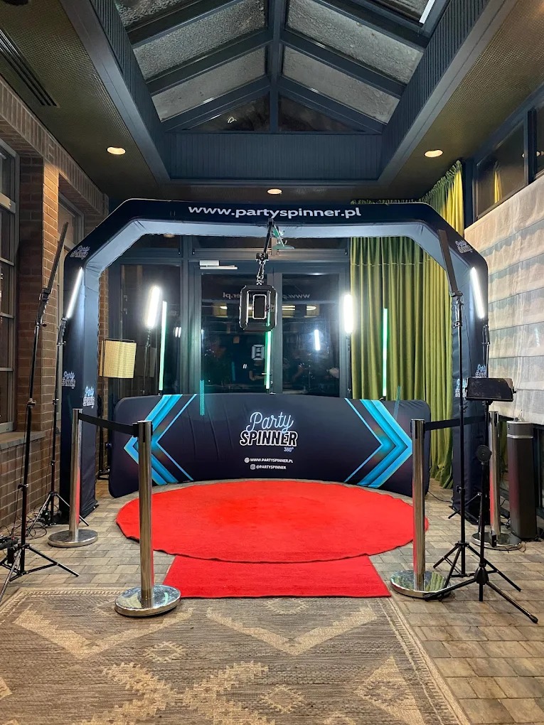 Gotowa do użycia fotobudka 360 z czerwonym dywanem i czarnym tłem z logo, oświetlona ledowymi lampami, gotowa na imprezę w eleganckim wnętrzu.
