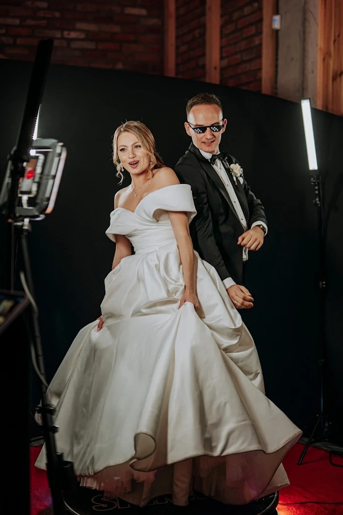 Para młoda pozuje w fotobudce: panna młoda w sukni z odkrytymi ramionami i pan młody w smokingu i pikselowych okularach, czarne tło i oświetlenie studyjne.