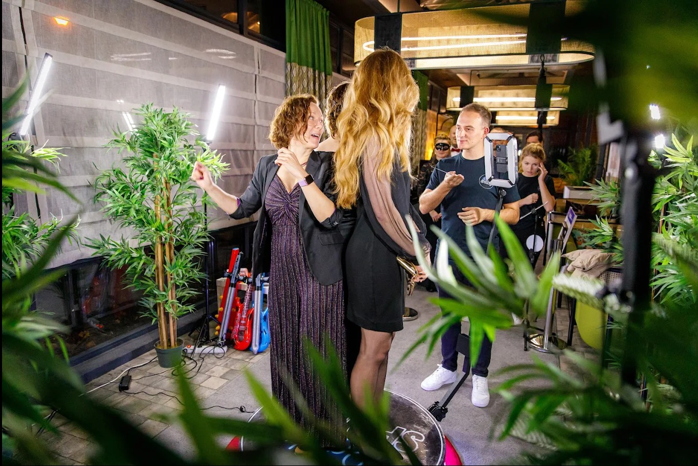 Dwie kobiety pozują do zdjęcia w fotobudce z rekwizytami. W tle widoczny operator i inne osoby, dekoracje roślinne i oświetlenie.