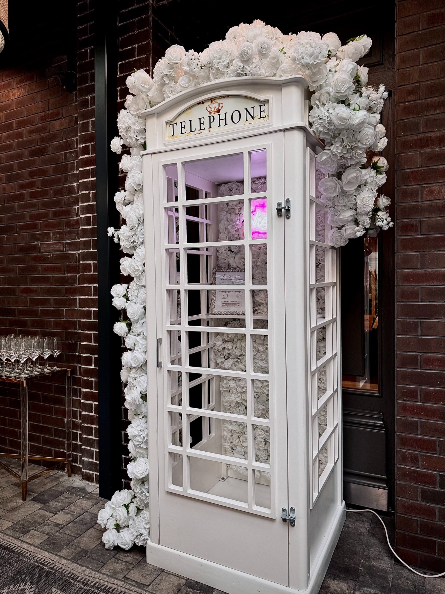 Biała budka telefoniczna, udekorowana białymi różami, ustawiona na tle ceglanej ściany. Wewnątrz widoczne oświetlenie i dekoracje kwiatowe.