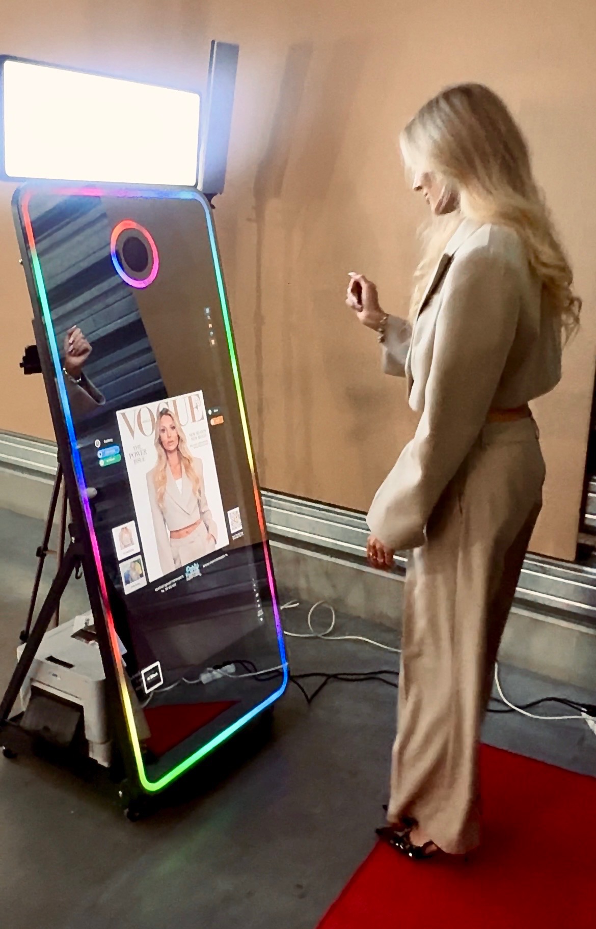 Kobieta w beżowym garniturze pozuje przed lustrzaną fotobudką z kolorowym oświetleniem LED, na czerwonym dywanie. Na ekranie fotobudki widoczny magazyn modowy.