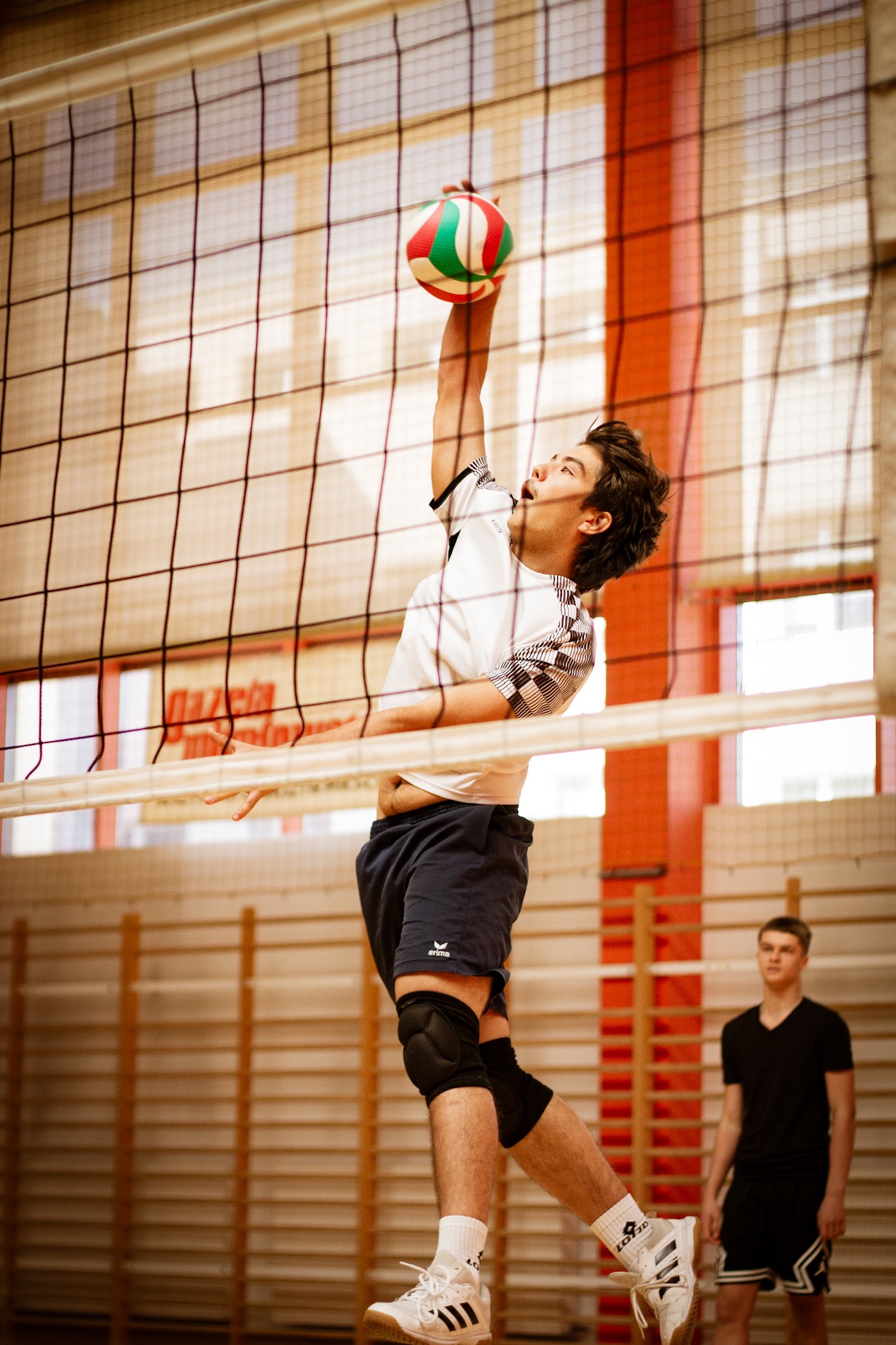 Młody siatkarz w białej koszulce z krótkim rękawem wykonuje atak przez siatkę. W tle drugi zawodnik i drabinki gimnastyczne.  Dynamiczne ujęcie z dołu.