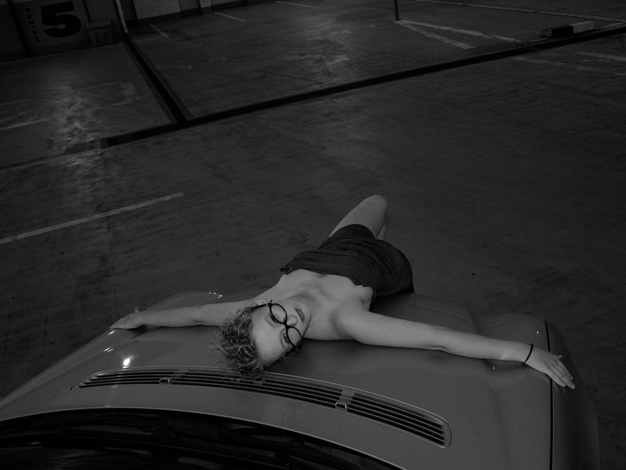 Sesja zdjęciowa w Czerwionce-Leszczynach: Młoda kobieta w okularach leży na masce samochodu na tle parkingu podziemnego. Nietypowe ujęcie w czerni i bieli.
