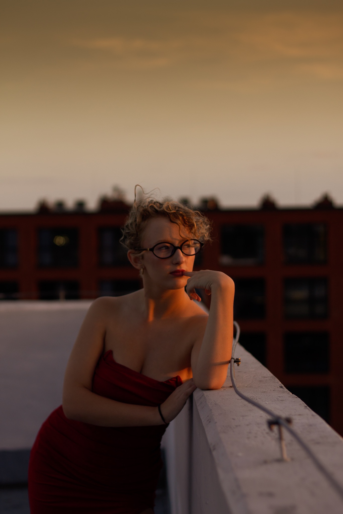 Portret kobiety w czerwonej sukience na tle zachodzącego słońca i budynku. Kobieta opiera się o balustradę i patrzy w bok, z palcem przy ustach.