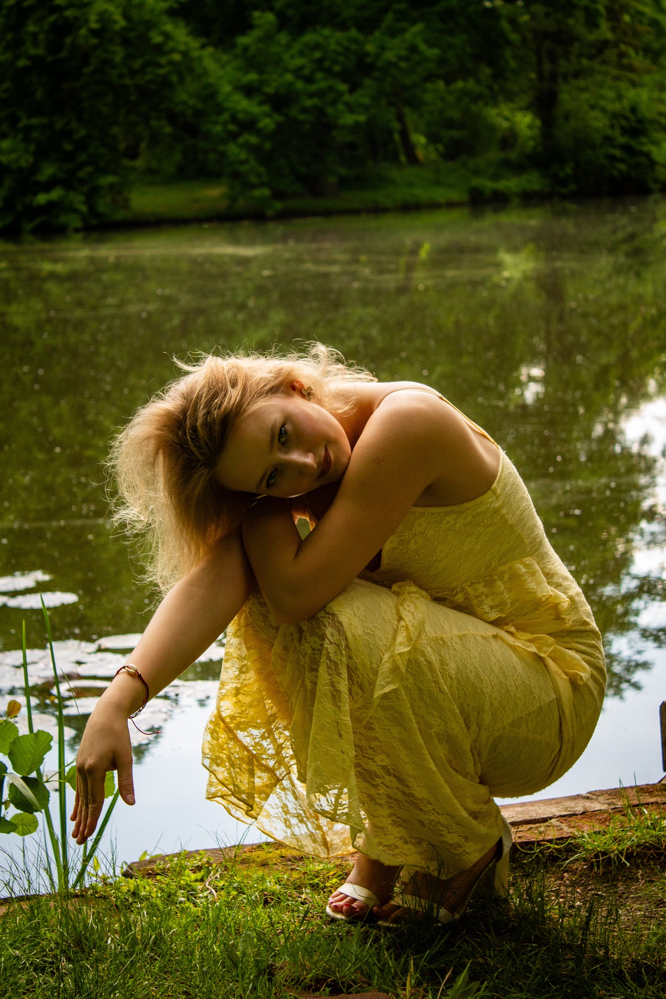 Portret młodej kobiety w żółtej sukience, skulonej na brzegu jeziora, z zamyślonym wyrazem twarzy i delikatnym uśmiechem.