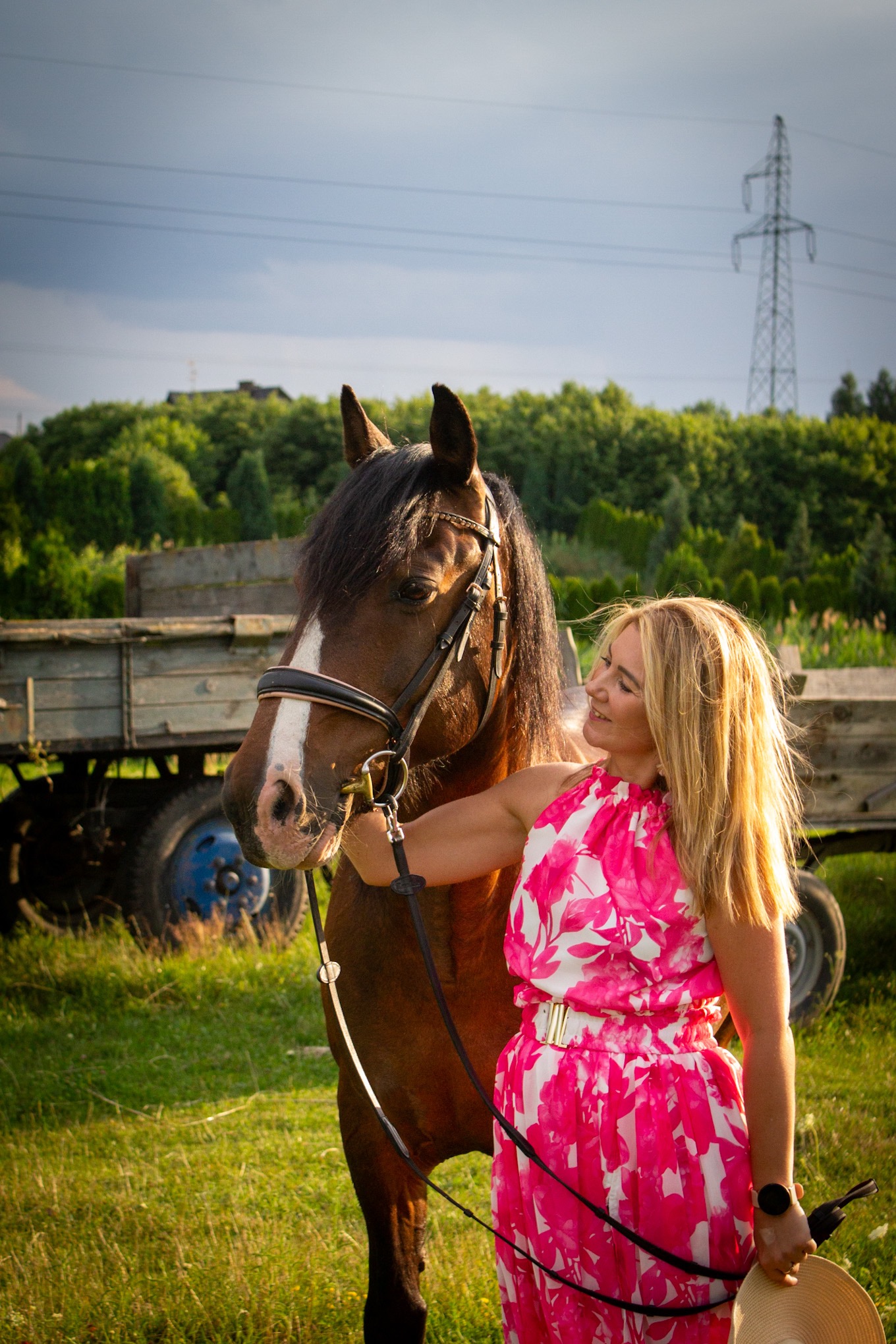 Kobieta w kwiecistej sukience z koniem na tle łąki i w oddali słupa energetycznego. Uśmiechnięta blondynka trzyma konia za uzdę. Sesja w plenerze.