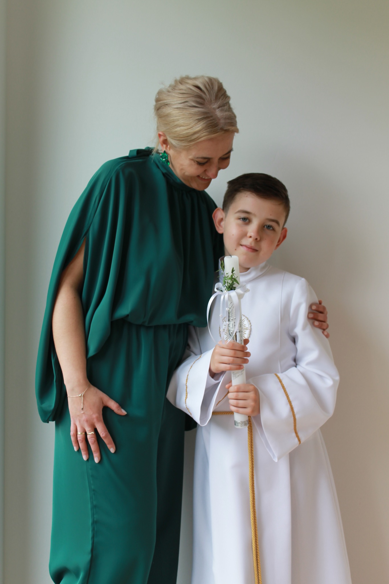 Chłopiec w albie komunijnej trzyma świecę, obok stoi uśmiechnięta mama w szmaragdowej sukience. Ujęcie portretowe, neutralne tło.