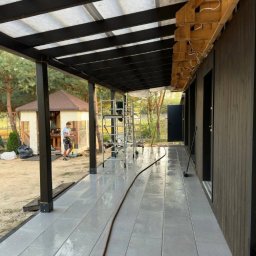 Dimex B.D.R - Zadaszony taras z szarą podłogą z płytek, czarne belki i słupy konstrukcyjne. W tle widoczna altana i osoba. Na tarasie stoi drabina.