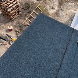 Dimex B.D.R - Granulowany dach z papy na deskowaniu, widok z góry, z fragmentem buta w rogu. Na tle palety, drabina i ziemia.