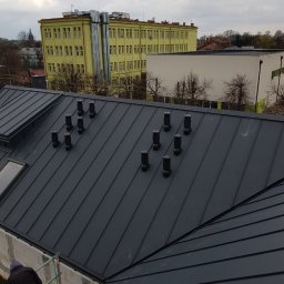 Dimex B.D.R - Nowoczesny, grafitowy dach z blachy na budynku w trakcie budowy, z widocznymi kominkami wentylacyjnymi i pracownikiem na rusztowaniu. W tle budynki i pochmurne niebo.