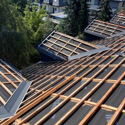 Dimex B.D.R - Konstrukcja dachu z łatami i membraną, widok z góry na tle zabudowy jednorodzinnej i zieleni, oświetlona słońcem.  Szczegółowo widoczne obróbki blacharskie.