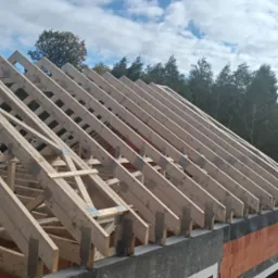 Konstrukcja więźby dachowej z drewna, zabezpieczona metalowymi łącznikami, na tle błękitnego nieba z chmurami. Widoczne elementy muru budynku z czerwonej cegły.