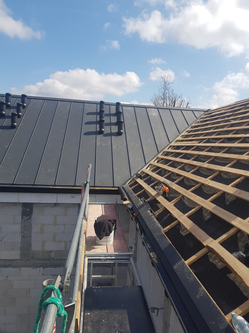 Dwa fragmenty dachu: jeden kryty blachą na rąbek stojący z odpowietrznikami, drugi w trakcie budowy z widoczną drewnianą konstrukcją. Praca na wysokości, rusztowanie.