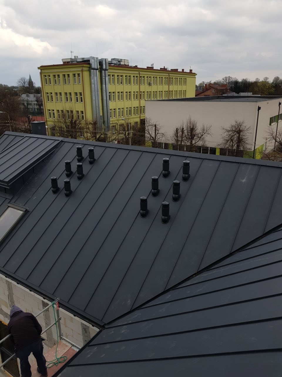 Nowoczesny, grafitowy dach z blachy na budynku w trakcie budowy, z widocznymi kominkami wentylacyjnymi i pracownikiem na rusztowaniu. W tle budynki i pochmurne niebo.
