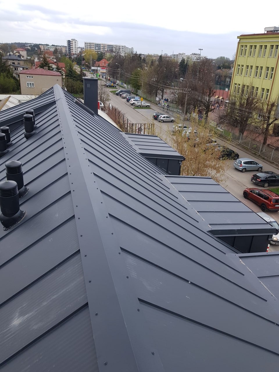 Szary dach z blachy na budynku mieszkalnym, widok z góry, kominki wentylacyjne, zabudowa miejska w tle.