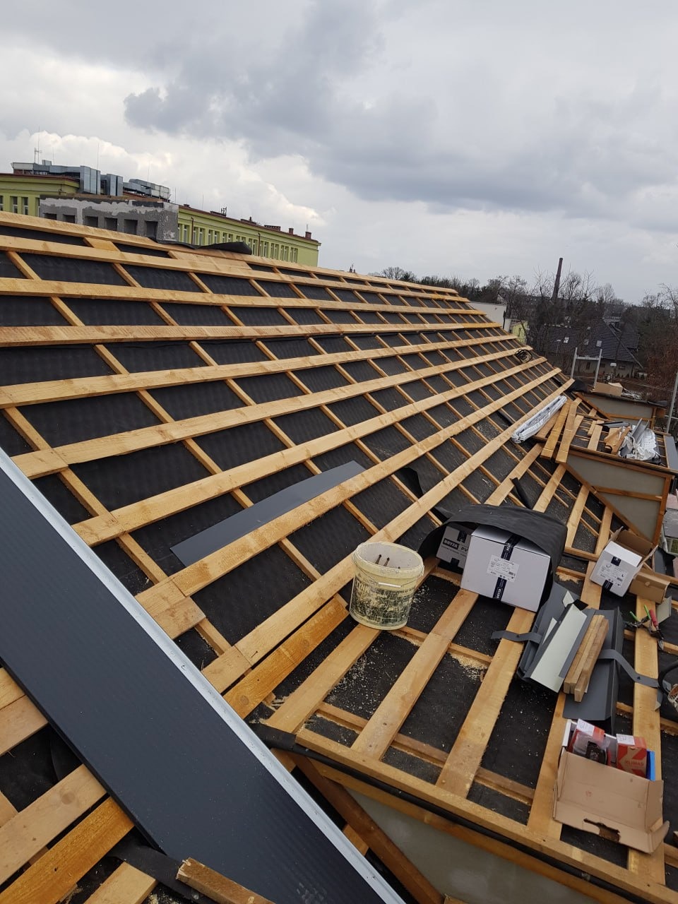 Dach w trakcie krycia: widoczna konstrukcja drewniana, czarna membrana, elementy blacharskie, narzędzia i materiały budowlane. W tle budynki miejskie pod pochmurnym niebem.