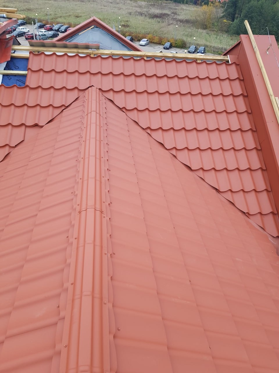 Czerwony dach z blachodachówki modułowej, widok z góry. Widoczne elementy konstrukcyjne i fragment budynku w tle. Dach o regularnej strukturze, z wyraźnymi liniami łączeń.