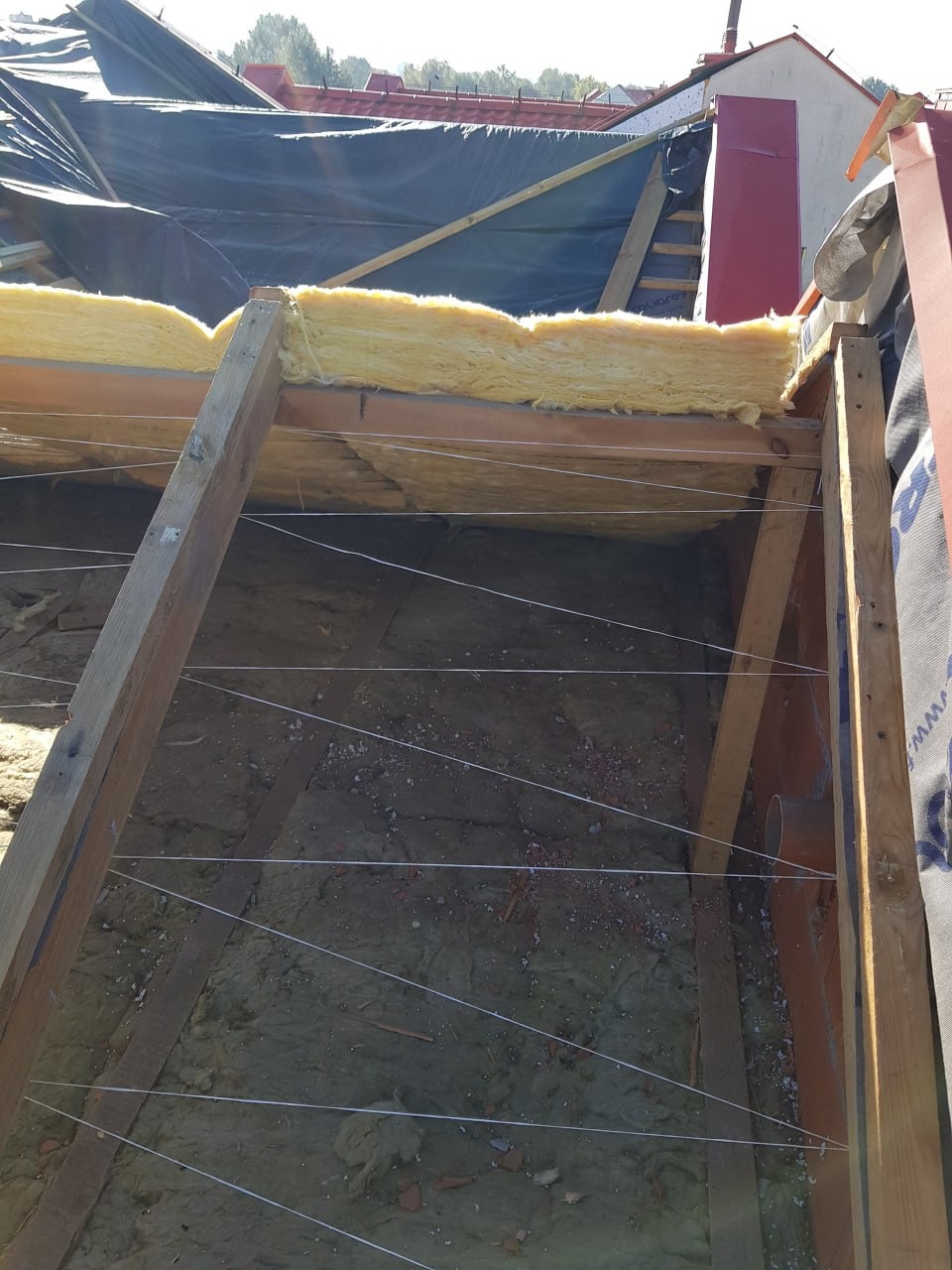 Poddasze w trakcie ocieplania: żółta wełna mineralna między krokwiami, zabezpieczona sznurkiem. Widoczne elementy konstrukcji dachu i fragment czarnej membrany dachowej.